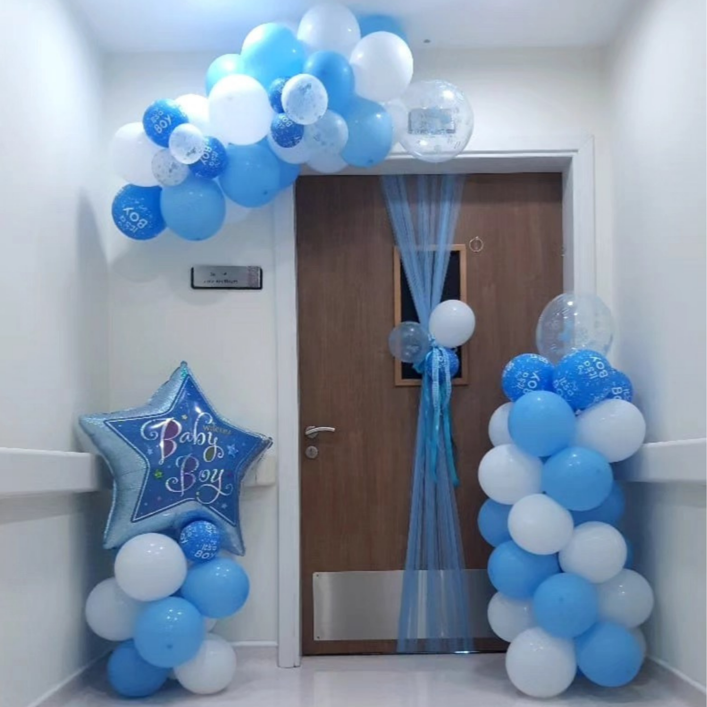 Decorative arch with blue and white balloons and a 'Baby Boy' star balloon standee outside a maternity room door.