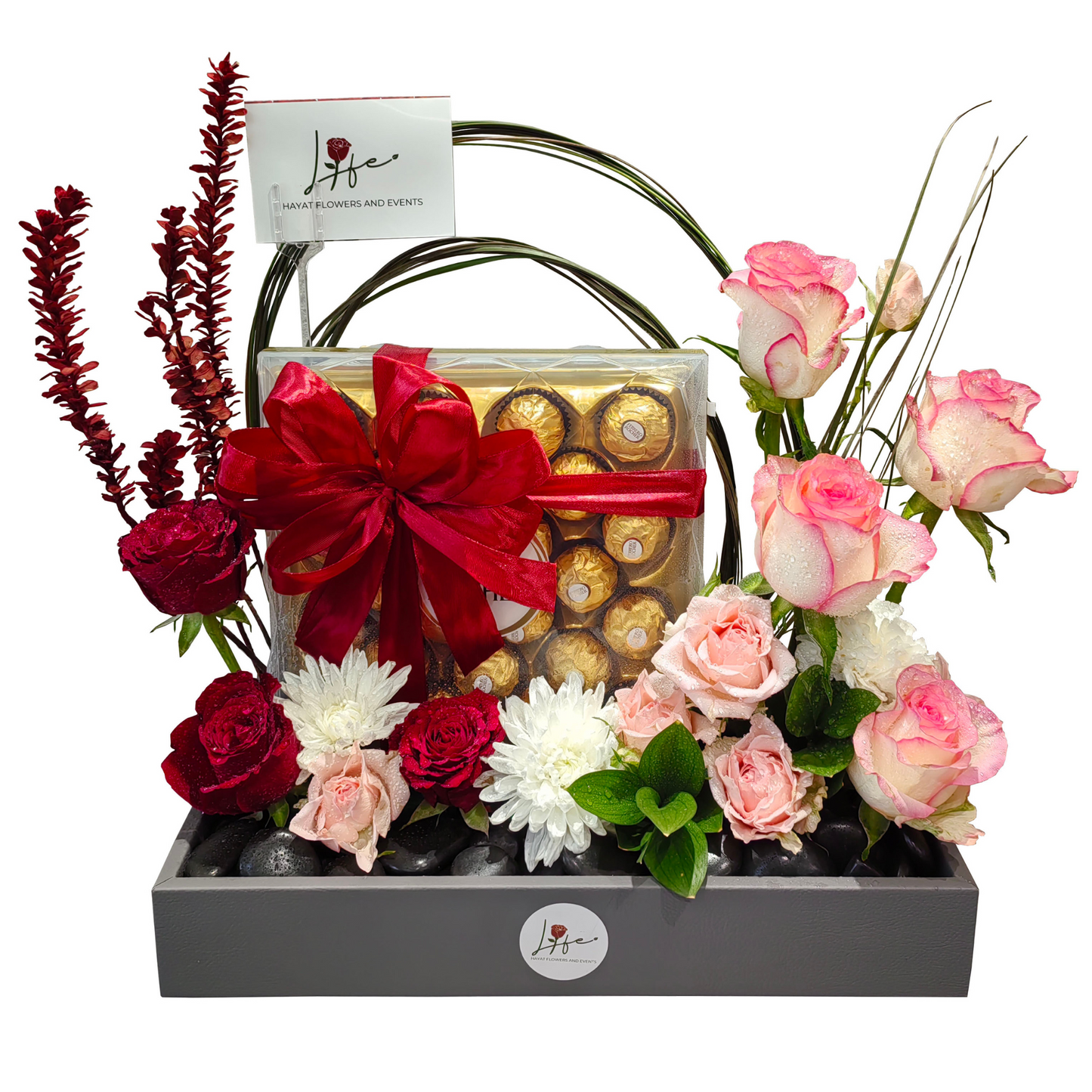 Floral arrangement with a box of chocolates, designed over black pebbles on a gray faux leather floral box.