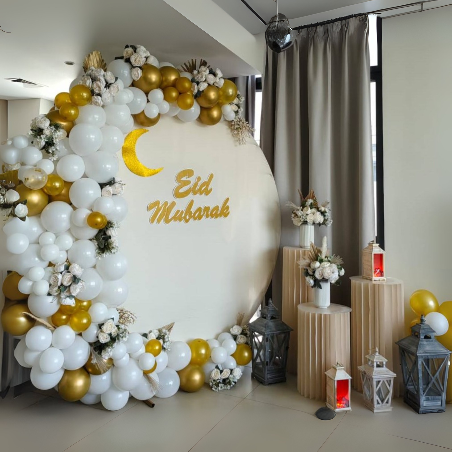 Decorative setup with 'Eid Mubarak' sign board, white and gold crescent moon balloons and floral arrangements. Decorative lamps on the floor, and faux flower arrangements in vases on top of three multi-level plinths.
