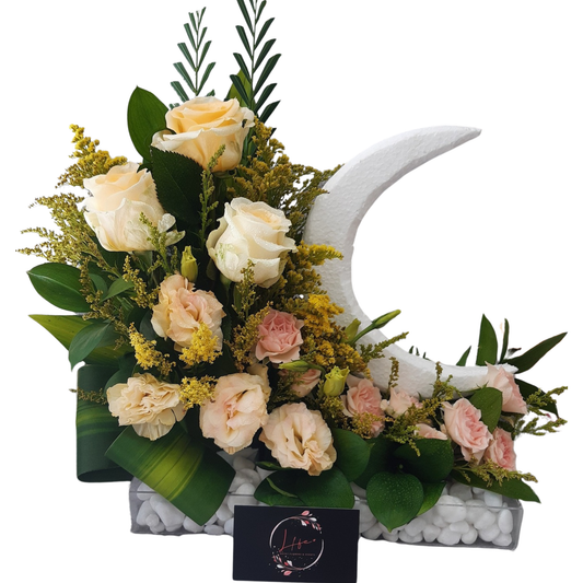 A crescent moon-inspired tabletop floral arrangement featuring peach and blush roses, lisianthus, golden solidago, and lush greenery, styled in a rectangular white pebble base — perfect for elegant home or event decor.