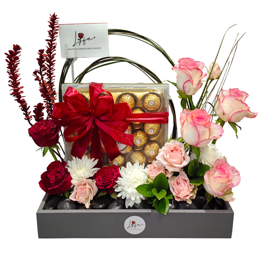 Floral arrangement with a box of chocolates, designed over black pebbles on a gray faux leather floral box.