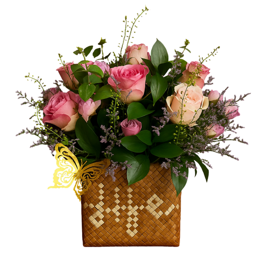 FFloral arrangement of pastel roses, ruscus leaves, and filler flowers in a handwoven 'Life' buri palm leaf box with optional gold butterfly accent by Hayat Flowers and Events. Solid white background.