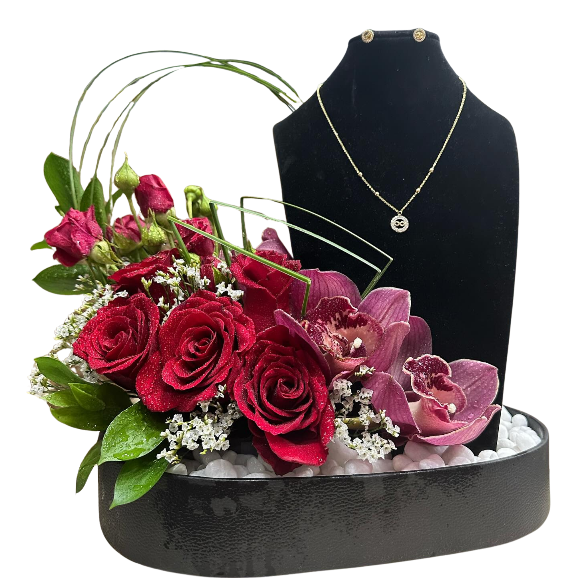 Elegant tabletop arrangement with red roses, pink cymbidium orchids, red lisianthus, white limoniums, greenery, and white pebbles on a black floristry tray, displayed beside an optional gold necklace and earring set on a velvet bust.