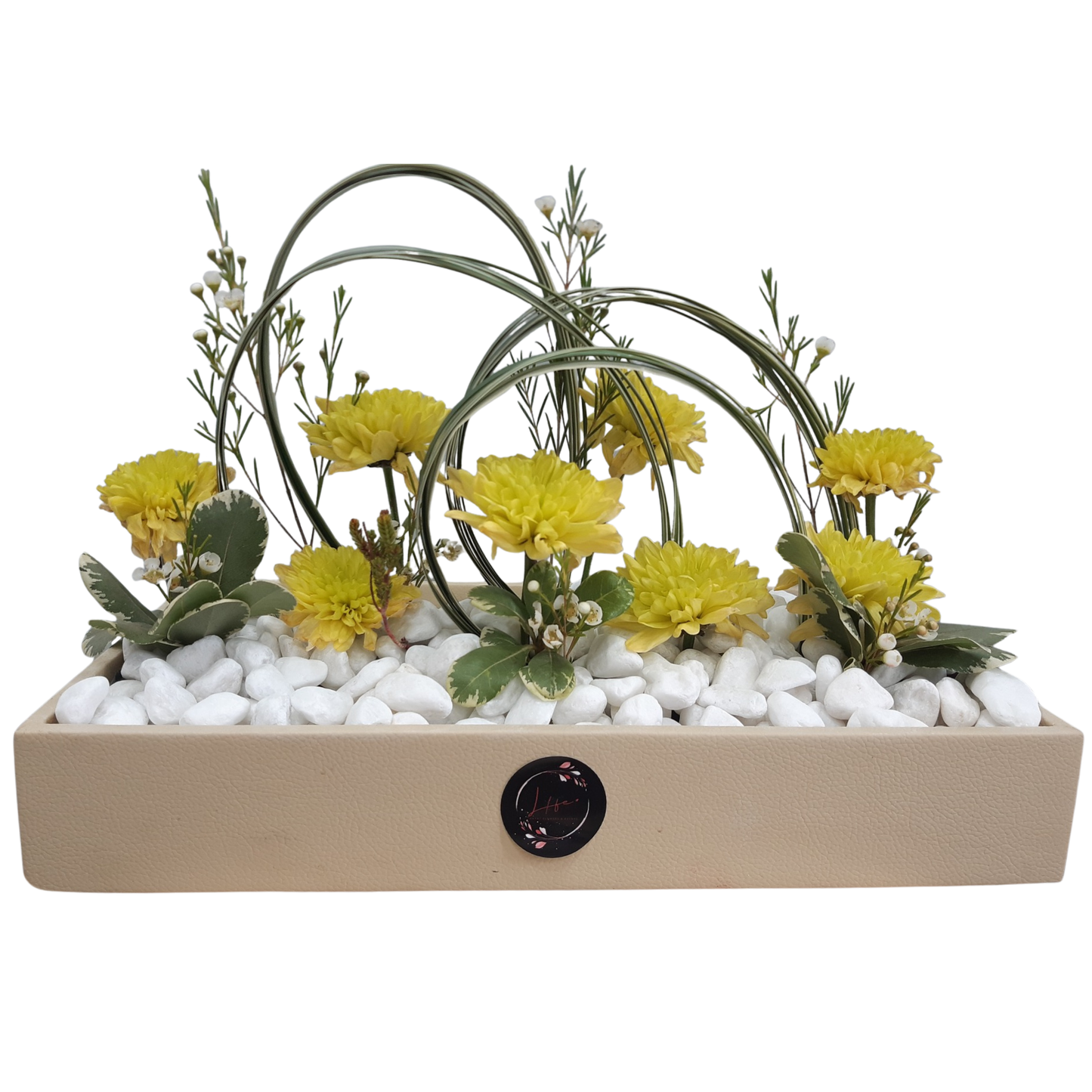 Minimalist tabletop floral arrangement featuring yellow flowers, white filler blooms, looped ornamental greens, and variegated foliage, set on white pebbles in a beige rectangular tray.
